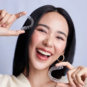 ผู้หญิงหัวเราะ ชูอุปกรณ์จัดฟันแบบใสไว้ข้างใบหน้า มือละชิ้น