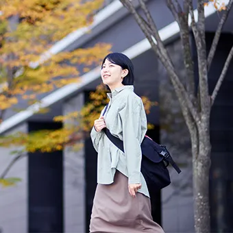 A smiling woman in a pale green jacket walking past autumn trees with a black bag worn across her body