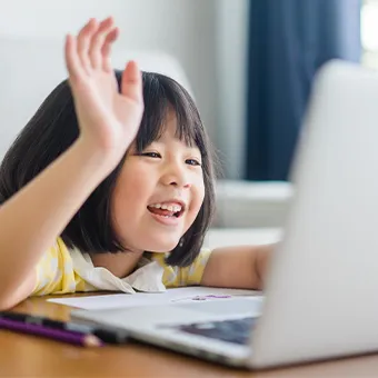 A smiling young girl in a yellow top raising her hand in front of a laptop