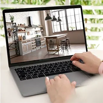 Hands on a laptop keyboard, the screen showing a kitchen with a wooden island and bar stools