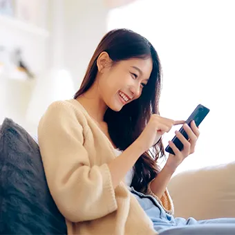 A smiling woman in a beige cardigan sitting on a sofa and tapping the screen of a phone