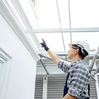 A man in a white hard hat and a checked shirt pointing up at the frame of a clear roof beside a white house