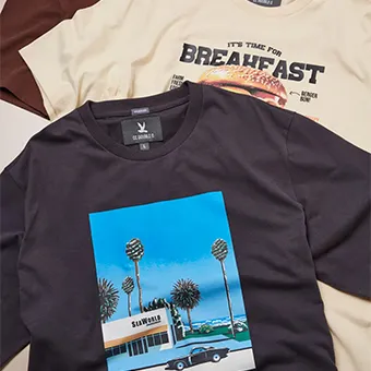 A black T-shirt printed with palm trees, a low building and a car, laid over a beige T-shirt with a breakfast print