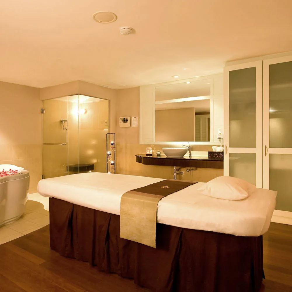 A treatment room with a massage bed made up in white and brown, a glass shower and a mirror over a basin
