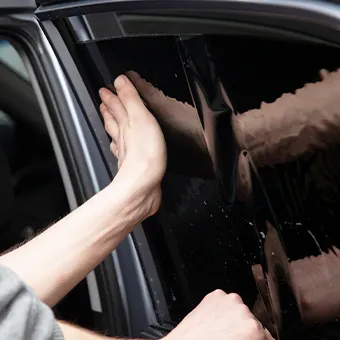 A hand smoothing a sheet of dark tint film onto a car side window