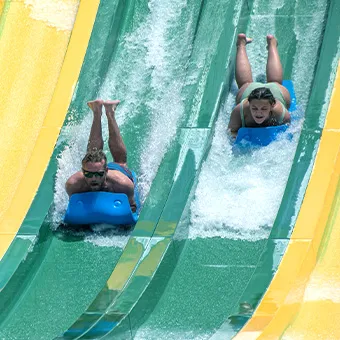 Two people lying on blue mats riding head first down a green racing slide