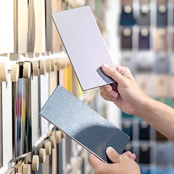Hands holding two metallic panel samples, one silver and one blue-grey, in front of a wall of colour samples