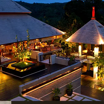 A lit wooden terrace and two pavilions with steep roofs at dusk, with forested hills behind