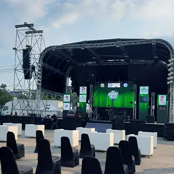 An outdoor stage under a curved truss roof, with green screens and speaker towers, behind white cube seats and black-covered chairs