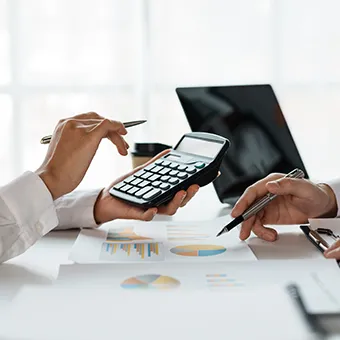 A hand holding up a calculator and a pen over printed charts on a desk as a second person points a pen at it, a laptop behind
