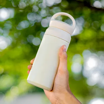 A hand holding a cream bottle with a loop handle against blurred green leaves