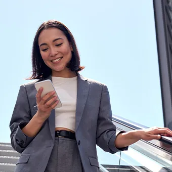 A smiling woman in a grey suit looking at her phone as she rides down an escalator