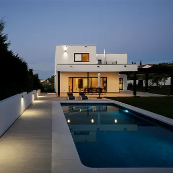 A white two-storey house lit up at night behind a swimming pool with underwater lights