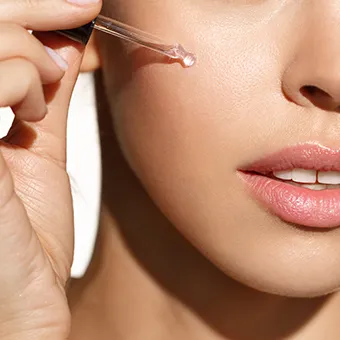 A hand holding a glass dropper with a drop of clear serum beside a woman’s cheek