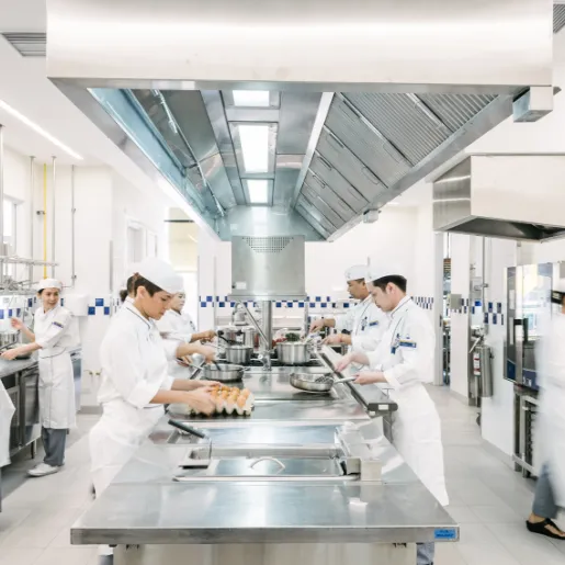 Cooks in white uniforms and caps working at steel stations under a large extractor hood in a bright kitchen