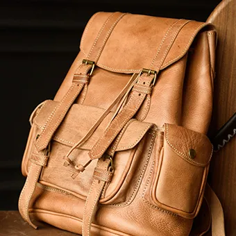 A light brown leather backpack with buckled straps and a front pocket against a wooden board