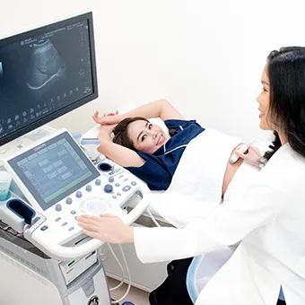 A woman in a white coat holds an ultrasound probe to the stomach of a smiling woman lying beside a scanner screen