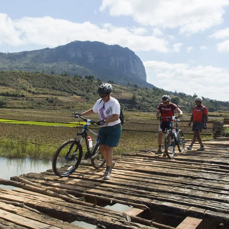 Three cyclists walking their mountain bikes across a wooden bridge over rice fields, a rocky mountain behind
