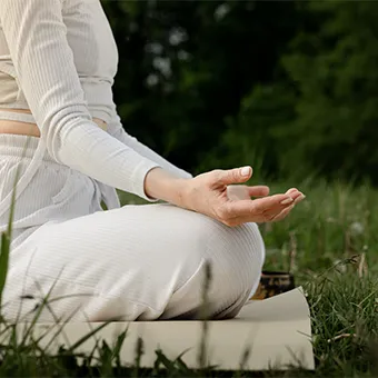 A woman in white sitting cross-legged on a mat on the grass, one hand resting palm up on her knee