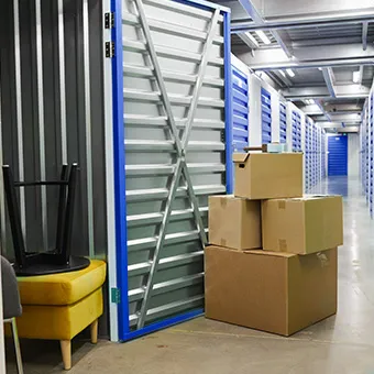 An open storage unit holding a yellow footstool and a black stool, with cardboard boxes stacked outside in a corridor of blue units