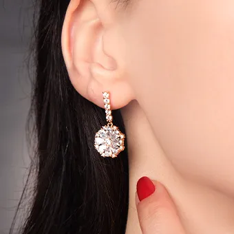 A drop earring with a large round clear stone in a rose gold coloured setting, worn by a woman with dark hair and red nails