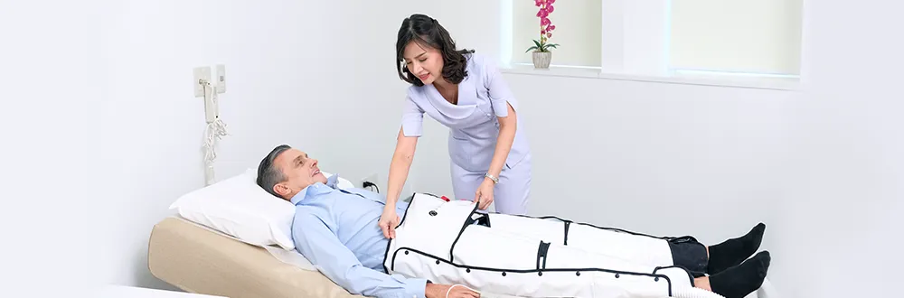 A woman in a lilac tunic smiles as she checks the padded white sleeves covering the legs of a man lying on a treatment bed