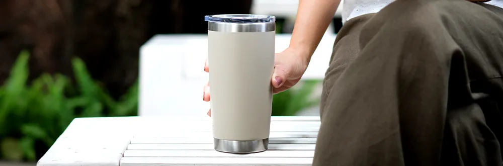 A hand holding a cream tumbler with a steel base and a clear lid on a white wooden bench, beside a person in olive trousers