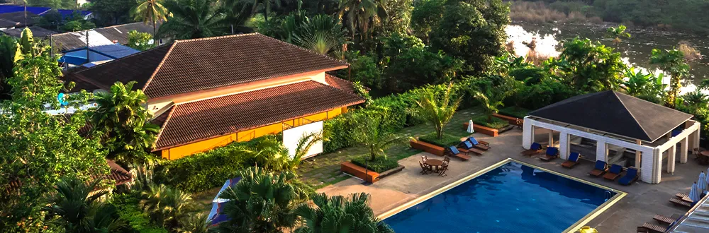 A swimming pool with blue sun loungers and an open pavilion beside a long building with a red tiled roof, among palm trees, seen from above