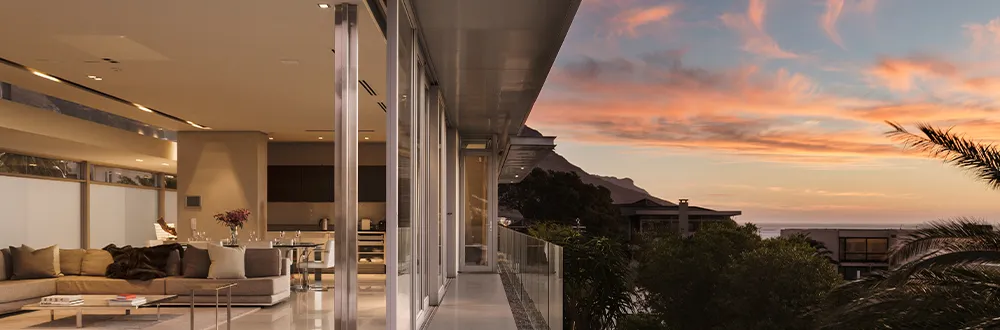A large lounge with cream sofas leading out to a glass balcony, with hills and the sea under a sunset sky