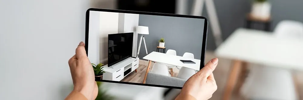 Hands holding up a tablet that shows a room with a white TV cabinet, a floor lamp and a white dining table with chairs