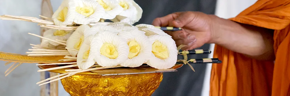 A hand in an orange monk’s robe reaching for white sandalwood flowers on a gold pedestal tray