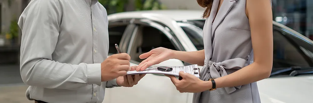 A man holding a pen and a clipboard with a car key on it, a woman’s hand reaching towards it, in front of a white car