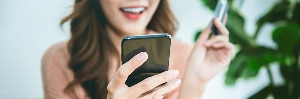 A smiling woman in a pink jumper holding a phone in one hand and a bank card in the other