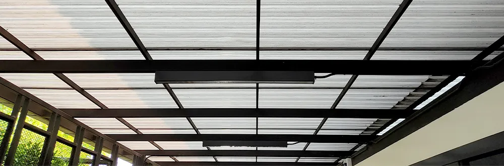 The underside of a roof of clear corrugated sheets on black steel beams beside a building, with 2 long lights fixed to the beams