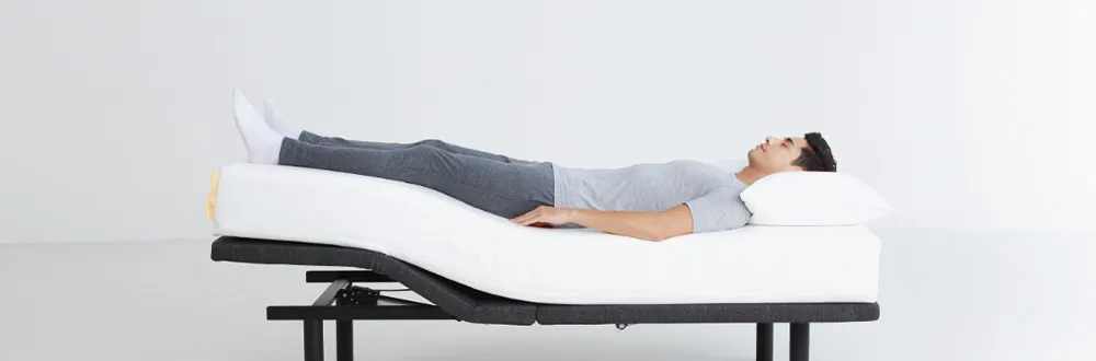 A man in grey lying on a white mattress on an adjustable bed base, with the head and knee sections raised