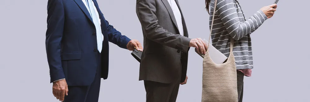 A man in a navy suit taking a wallet from the man in front, who is taking a notebook from a woman’s woven shoulder bag while she looks at her phone