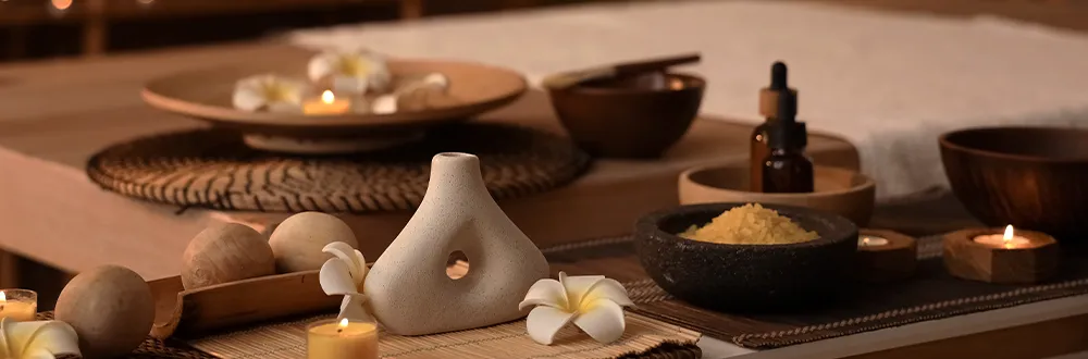 A spa table set with a white ceramic vase, frangipani flowers, lit candles, a small amber dropper bottle and wooden bowls, one holding yellow salt