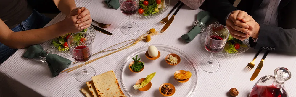 A table laid with a white cloth, wine glasses, salads and a glass plate of small bites, two diners’ clasped hands on either side