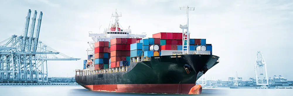 A container ship stacked with coloured containers sailing away from a port, with cranes behind it