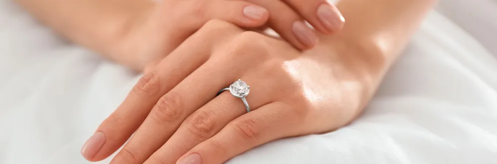 A woman’s hands resting on white fabric, a ring with a single round stone on one finger