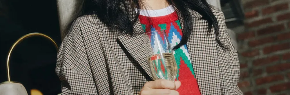A woman with long dark hair in a brown check blazer over a red, green and blue patterned knit jumper, holding a glass of sparkling wine beside a brick wall