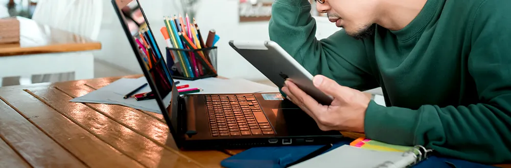 A man in a green jumper looking at a tablet beside an open laptop on a wooden table, with a pot of coloured pencils and a ring binder