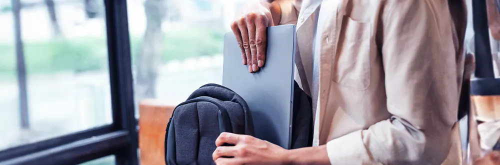 A person in a beige shirt sliding a silver laptop into a dark grey backpack beside a window