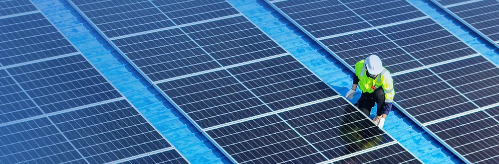 A worker in a hard hat and a high-visibility vest kneeling among rows of solar panels on a blue roof