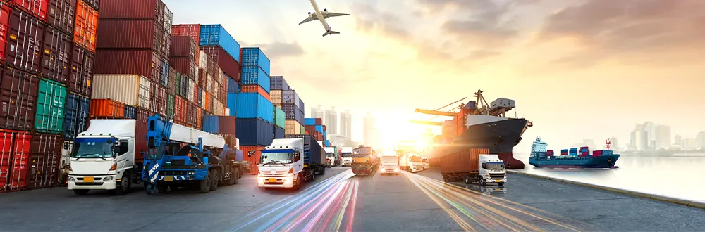 Lorries and a crane truck on a port road between stacks of shipping containers, with container ships, a plane overhead and a city skyline at sunset