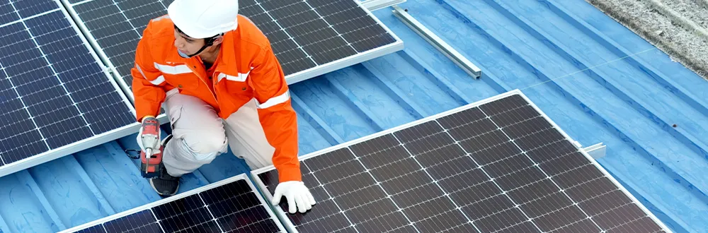 A worker in an orange uniform and a white hard hat kneeling between solar panels on a blue metal roof and holding a power drill