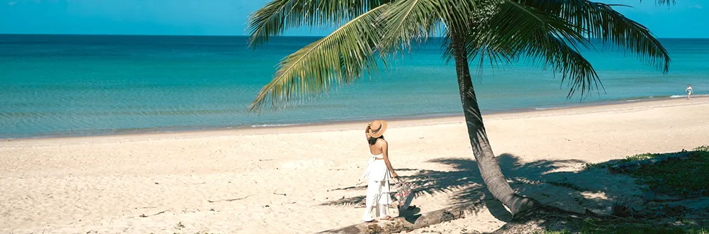A woman in a sun hat and white clothes standing under a palm tree on a white sand beach beside a turquoise sea