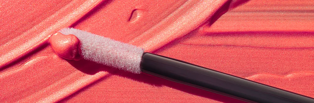แปรงหัวลิปกลอสสีขาววางพาดอยู่บนรอยลิปกลอสสีชมพูอมส้ม
