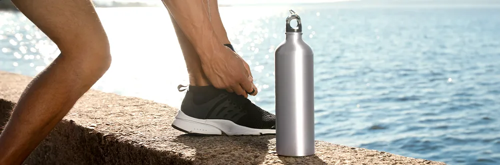 A runner tying a shoelace on a stone ledge by the sea, a silver metal water bottle standing beside the shoe