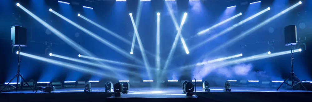 Beams of white and blue light crossing above an empty stage, with stage lights along the front edge and a loudspeaker on a stand at each side